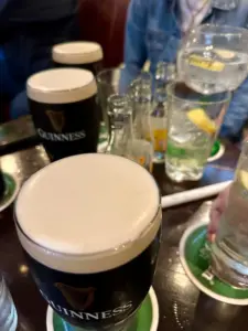 Two Guinness pint glasses with creamy heads sit on a table among clear glasses and bottles with lemon slices. People in casual clothing are partially visible in the background, evoking the lively atmosphere of a pub.