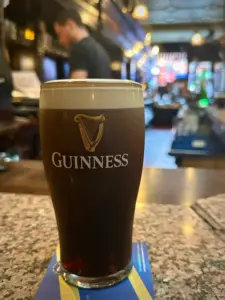 A Guinness pint glass sits on a bar counter, with a creamy head and the Guinness logo on the glass. The background reveals a softly focused bartender and a warmly lit pub interior.