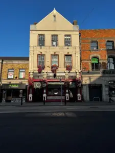 A three-story pub with hanging flower baskets and red trim stands between two brick buildings under a clear blue sky, its sign featuring a classic Guinness pint glass on a quiet city street.