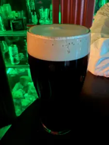 A pint glass filled with stout topped with a creamy foam head sits on a black surface, with green-lit shelves displaying various objects in the blurry background.