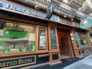 The exterior of a pub called "The Holy Cross" with signs for Guinness and Spaten beers, large windows displaying bottles, and a wooden entrance on a city street.