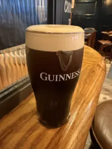 A Guinness pint glass with a creamy head sits on a polished wooden bar in a cozy pub setting, with empty chairs and tables visible in the background.