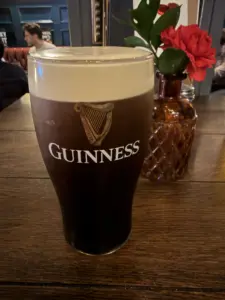 A Guinness pint glass with a creamy head sits on a wooden table in a pub; behind it is a decorative vase with red flowers and green leaves.