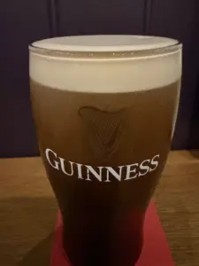 A Guinness pint glass with a creamy head sits on a red coaster atop a wooden surface in a pub-like setting, against a dark background.
