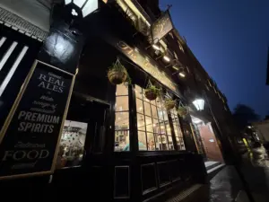 A warmly lit pub with large windows and hanging plants is seen from a low angle at dusk. Signs advertise a Guinness pint glass, premium spirits, and food. The street outside glistens from the rain.