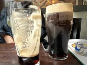 Two pints of dark beer sit on a wooden table; one is half-finished with foam residue, the other is full. A phone and magazine are nearby, and a person is partially visible in the background.