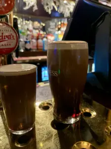 Two pints in Guinness pint glasses sit on a brass bar counter at a pub, with taps and blurred bottles visible in the background.