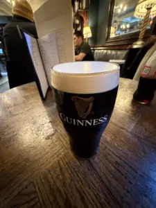 A pint of Guinness stout sits on a wooden pub table with a foam head. A menu, another drink, and people are visible in the background, along with warm lighting and decorative mirrors.