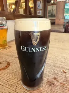 A Guinness pint glass with a creamy head sits on a wooden table in a cozy pub, with another glass of cider and classic pub decor visible in the background.
