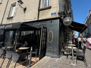 A street view of an Irish pub with outdoor seating on a cobblestone sidewalk; people sit at tables under a black awning. The pub is painted dark with gold accents, and a Guinness pint glass sign hangs above the entrance.