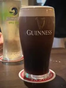 A pint of Guinness stout with a creamy head sits on a coaster in the foreground, while a pint of Rockshore Apple Cider stands in the background on a wooden table.
