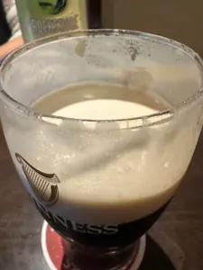 A close-up of a partially full glass of Guinness stout beer with creamy foam on top, sitting on a coaster on a wooden table. A bottle of Rockshore is blurred in the background.
