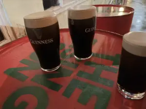 Three Guinness pint glasses with creamy heads sit on a large red table at a pub. The scene appears to be outdoors, with pavement and building walls in the background.