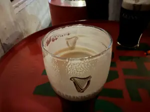 A partially finished Guinness pint glass sits on a red table, with foam clinging to the inside of the glass. Another pint is visible in the background, and the table features green and red markings, evoking a lively pub scene.
