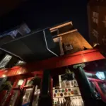 Nighttime view looking up at a red-bricked pub with a green awning and hanging plants, warmly lit. The sign reads "THE RIS," with various colorful signs, including a Guinness pint glass, visible in the window.