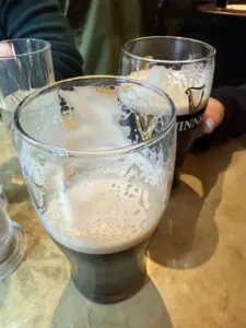 Two partially-drunk Guinness pint glasses sit on a table at the pub, with foam residue clinging to the insides. Another glass and two people’s hands are visible in the background.