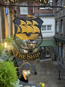A decorative pub sign with a golden ship and waves hangs above a narrow alley, reading "THE SHIP" in gold letters. A Guinness pint glass is painted on the sign. The wet street below hints at recent rain, with greenery in the foreground.