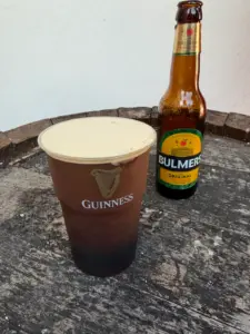 A plastic Guinness pint filled with dark stout topped with a creamy head sits on a weathered wooden surface, next to a glass bottle of Bulmers Original Irish Cider.