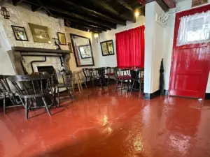 A cozy pub interior with wooden chairs and tables, a white brick fireplace, framed pictures on the walls, and red curtains and door, all set on a shiny red floor.