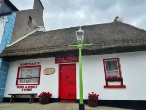 A quaint white cottage with a thatched roof, red door, and red-trimmed windows, housing “Frank Dorrian’s Wine & Spirit Store.” A green lamp post stands in front, with flower boxes under the windows.