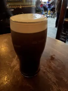A Guinness pint glass with a creamy white head sits on a wooden table in a dimly lit pub, with blurred people and chairs visible in the background.