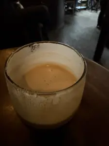 A close-up of a nearly empty Guinness pint glass with creamy foam clinging to the inside, sitting on a wooden table in a dimly lit pub. The background is dark and out of focus.