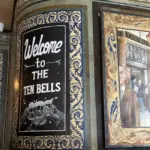 A decorative tiled wall features a black sign reading "Welcome to The Ten Bells" with an illustration of clinking Guinness pint glasses, next to a framed painting showing people outside a building.