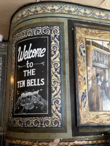 A decorative tiled wall features a black sign reading "Welcome to The Ten Bells" with an illustration of clinking Guinness pint glasses, next to a framed painting showing people outside a building.