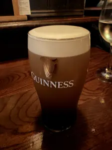 A Guinness pint glass with a creamy head sits on a wooden pub counter, next to a partially visible wine glass in the background.