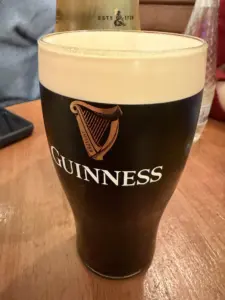 A pint glass filled with Guinness stout, showing a creamy head and the Guinness logo with a harp, sits on a wooden table. Other drinks and a mobile phone are visible in the blurred background.