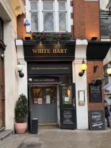 Entrance to the White Hart pub in London, with a black and gold sign, flowers above the door, lantern-style lights, and a menu displayed outside. A potted plant sits beside the door on a cobblestone street.