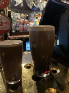 Two pints in Guinness pint glasses, one large and one small, rest on a brass bar counter in a dimly lit pub with various liquor bottles and taps blurred in the background.