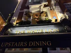 Nighttime view of The White Lion pub exterior, with illuminated gold lettering and flower baskets on the balcony above. A Guinness pint glass glows in the window, while a sign advertises "Upstairs Dining.
