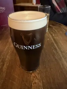 A pint of Guinness stout with a creamy head sits on a wooden table in a cozy pub, with a glass of light-colored drink and a blurred person in the background.