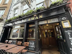 The exterior of The White Swan pub, with black painted facade, gold lettering, outdoor seating, and signs advertising fish and chips and live sports. Plants line the awning above the entrance.