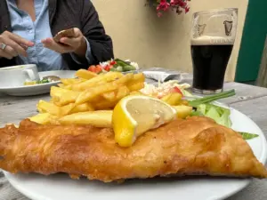 A plate of fish and chips with a lemon wedge, fries, and salad sits on a wooden table. A pint of Guinness and another person eating are visible in the background.