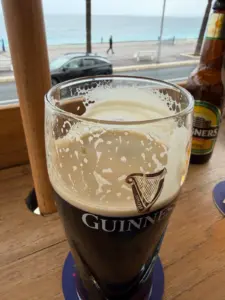 A partially drunk pint of Guinness on a wooden table, with a bottle of Magners cider nearby. In the background, a road, railing, and the sea are visible on an overcast day.