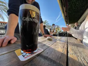 A close-up of a pint of Guinness on a wooden outdoor table in sunlight, with people sitting and talking, palm trees and blue sky in the background, and another drink and coaster visible.