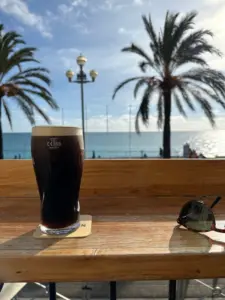 A pint of dark beer sits on a wooden table outside, next to sunglasses, with palm trees, a promenade, and the ocean visible under a sunny sky in the background.