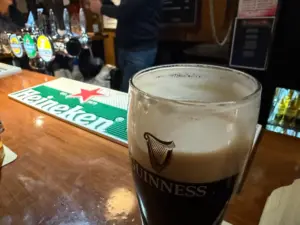 A pint of Guinness sits on a wooden bar counter near a Heineken-branded mat, with beer taps and a bartender in the background. The scene is warmly lit, creating a cozy pub atmosphere.