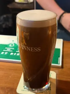 A pint glass of Guinness stout with a creamy head sits on a coaster atop a wooden bar counter, with a blurred hand and green bar mat in the background.