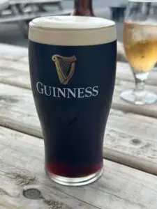 A pint of Guinness stout with a creamy head sits in a Guinness pint glass on a wooden outdoor table at a pub. In the background, there's another glass and more picnic tables.