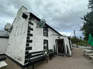 A white, two-story guinness pint glass pub with black trim, a Heineken sign, benches outside, potted plants by the entrance, and cloudy skies overhead. A road and green trees are visible in the background.