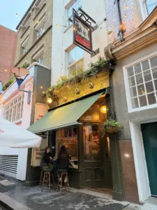 Two people sit at a small table outside "Ye Olde White Horse," a cozy pub with a green awning and warm lights. The pub is on a narrow street with flower boxes above the entrance and buildings close together.
