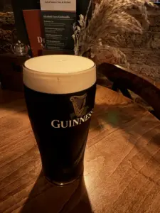 A pint of Guinness with a creamy head sits on a wooden table in a cozy pub, with menus and dried plants in the background.