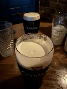 Two pints of Guinness with creamy heads sit on a wooden table in a dimly lit pub, alongside an empty pint glass and a textured tumbler. A brick wall and part of a blue door are in the background.