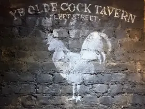 White painted rooster and the words “Ye Olde Cock Tavern - Fleet Street -” appear on a rough, dark brick wall.