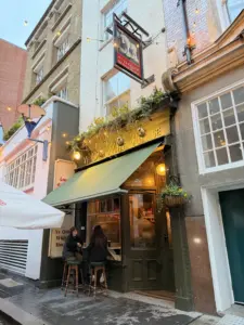 Two people sit on stools outside "Ye Olde White Horse" pub with a green awning, warm lights, and hanging plants. A Guinness pint glass rests nearby. It's dusk, with the street lined by tall buildings and string lights above.