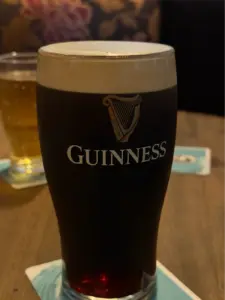 A pint glass of Guinness stout with a creamy head sits on a coaster on a wooden table, with a lighter drink in a smaller glass in the background.