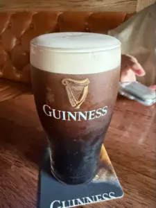A pint of Guinness stout with a creamy head sits on a branded coaster on a wooden table, with a tufted leather bench and a blurry hand in the background.
